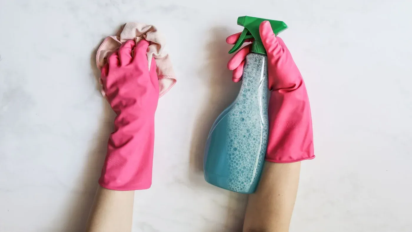 Händer i rosa gummihandskar håller en trasa och en sprayflaska med blå tvållösning mot en ljus marmorskiva