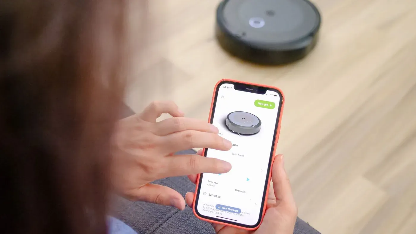Hand som håller en mobil med en robotdammsugares app, med roboten suddig i bakgrunden
