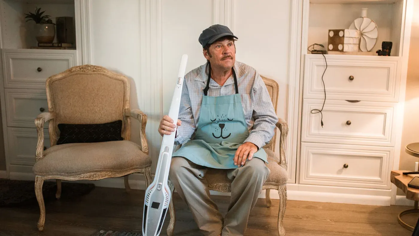 Man i förkläde sitter i en fåtölj med en vit sladdlös skaftdammsugare